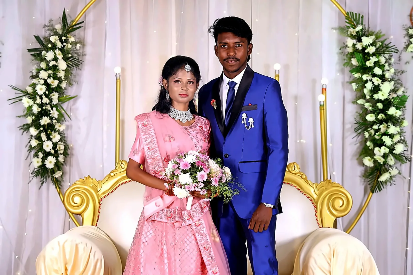 Bride and groom seated on a golden sofa under a pastel floral arch at a Chennai wedding reception stage
